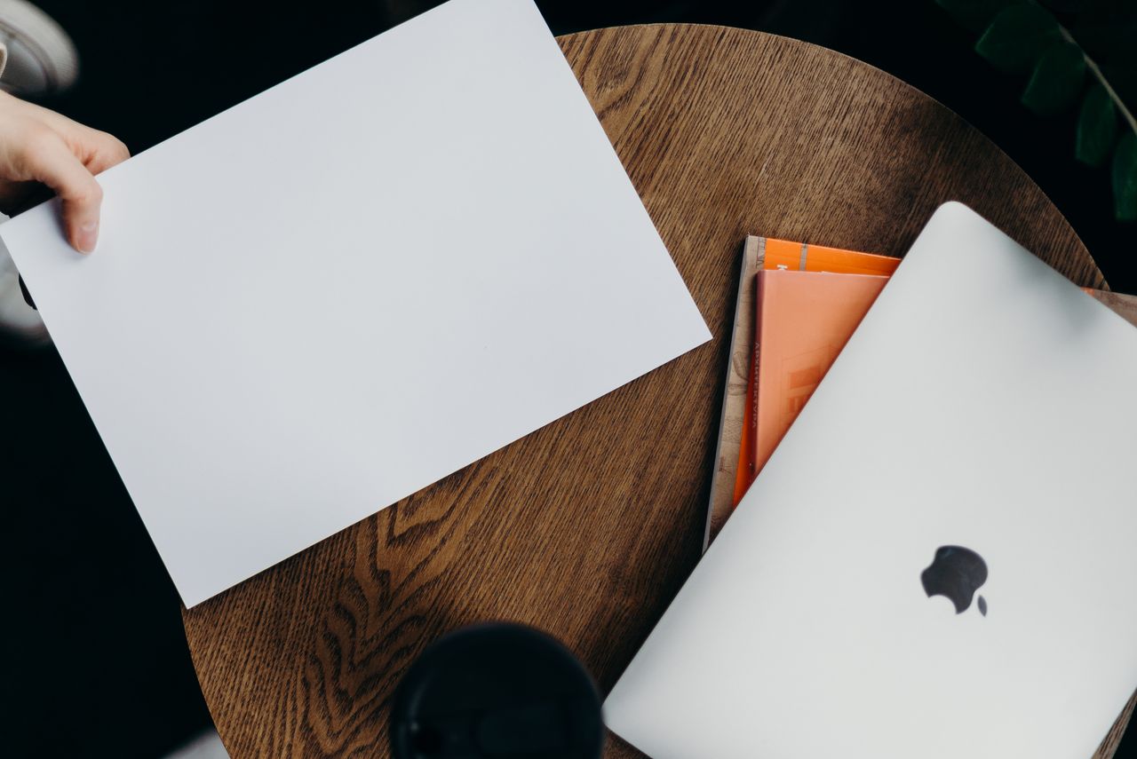 who-we-are A top-down view of a minimalist workspace featuring a laptop and blank paper on a wooden table.